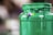 A close-up view of a green plastic jar with paint oozing from the top. The jar is prominently focused, and there are small patches of purple and yellow on the surface. The background is blurred and has a mix of brown and green tones, indicating an indoor setting.