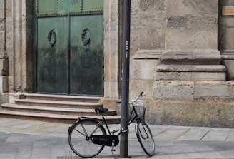 A black bicycle is securely locked to a grey metal pole on a stone pavement. The bicycle is positioned in front of an old, large door with a greenish patina and ornate details, set in a stone wall. The wall and pavement have a weathered and historic appearance.