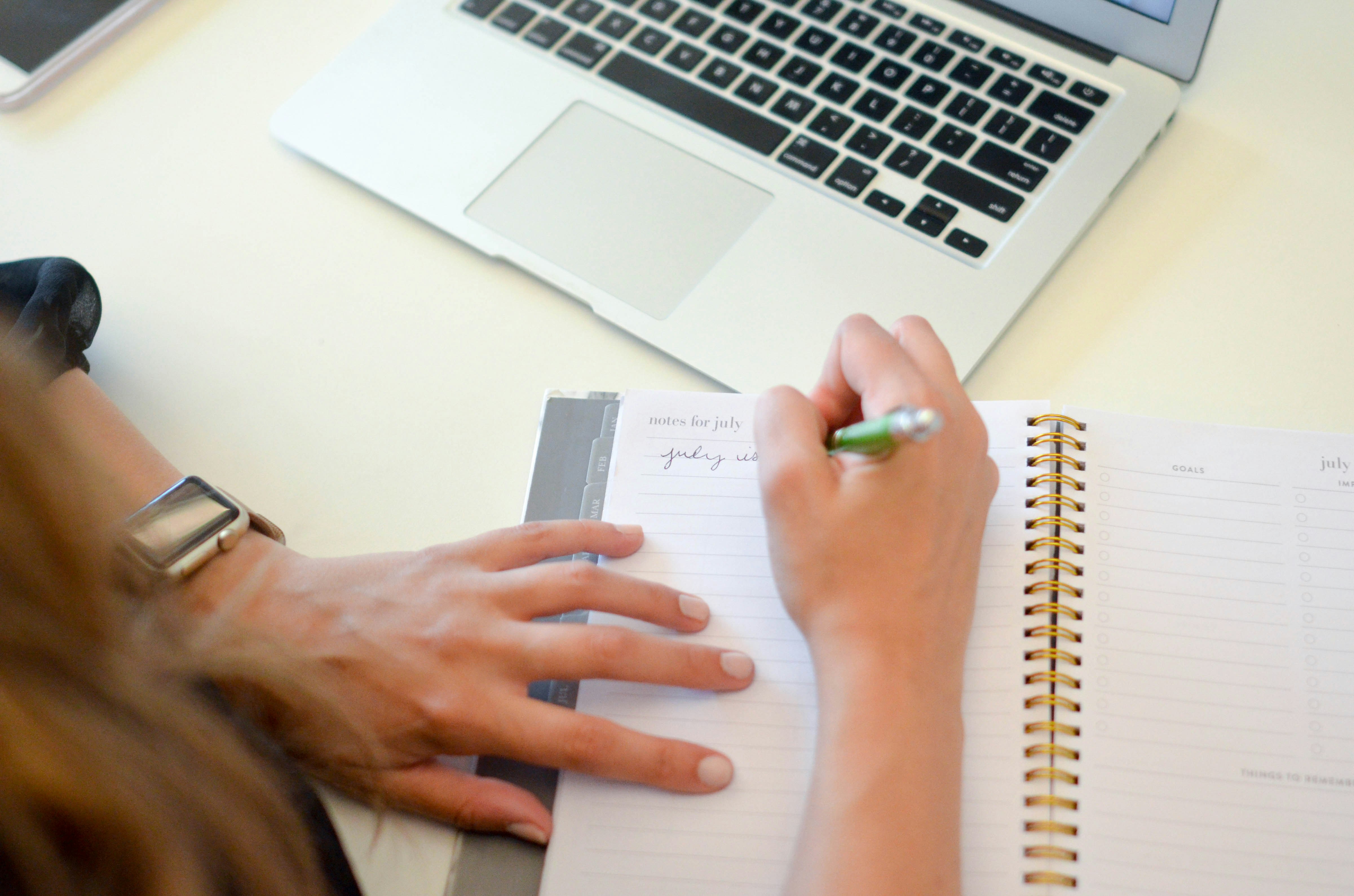 Person writing an article in a notebook beside a laptop
