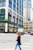 A city street intersection featuring tall buildings with large windows and ornate architecture. A green street sign labeled 'Woodward' is visible. There is a 'WEWORK' location on the corner at street level. Various vehicles, including a red car and a vintage-themed truck, are on the street. A person is crossing the street in the foreground, blurred due to motion.