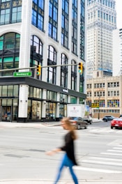 A city street intersection featuring tall buildings with large windows and ornate architecture. A green street sign labeled 'Woodward' is visible. There is a 'WEWORK' location on the corner at street level. Various vehicles, including a red car and a vintage-themed truck, are on the street. A person is crossing the street in the foreground, blurred due to motion.