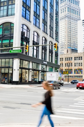 A city street intersection featuring tall buildings with large windows and ornate architecture. A green street sign labeled 'Woodward' is visible. There is a 'WEWORK' location on the corner at street level. Various vehicles, including a red car and a vintage-themed truck, are on the street. A person is crossing the street in the foreground, blurred due to motion.