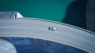 An aerial view of a curved dam structure with a single vehicle driving on the road on top. The water body adjacent to the dam is a bright turquoise color, contrasting with the gray concrete of the dam and road.