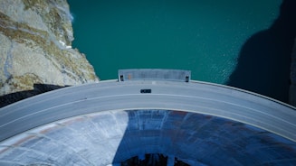 Aerial view of a large, curved concrete dam with a roadway on top, spanning across a turquoise body of water. A single vehicle is driving along the road, and the surrounding landscape features rocky, arid terrain.