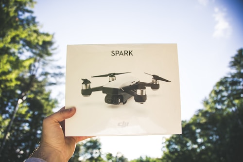 A hand holding a box with an image of a drone labeled 'SPARK' against a backdrop of clear blue sky and green trees.