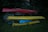 Three kayaks are stacked on a wooden rack in a grassy area surrounded by dense greenery. The kayaks are in different colors: red, yellow, and blue. The lighting is dim, suggesting that it might be early morning or late evening.