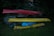 Three kayaks are stacked on a wooden rack in a grassy area surrounded by dense greenery. The kayaks are in different colors: red, yellow, and blue. The lighting is dim, suggesting that it might be early morning or late evening.