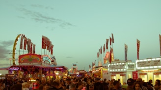 A bustling carnival scene with colorful rides, bright lights, and numerous flags waving. Many people are gathering, creating a lively atmosphere. Ticket booths are visible in the foreground, advertising various pricing options. The sky is clear with a hint of early evening light.