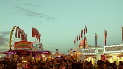 A bustling carnival scene with colorful rides, bright lights, and numerous flags waving. Many people are gathering, creating a lively atmosphere. Ticket booths are visible in the foreground, advertising various pricing options. The sky is clear with a hint of early evening light.