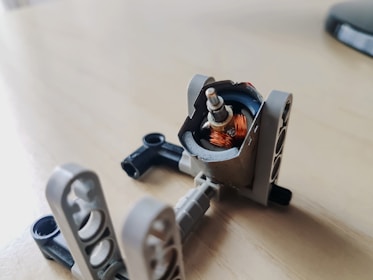 A small, partially disassembled electric motor with visible copper coils is placed on a light wooden surface. Surrounding the motor are gray plastic components that appear to be part of a building block kit.