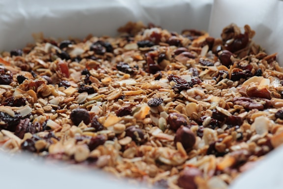 A close-up view of a mixture of granola consisting of oats, nuts, seeds, dried fruits, and possibly coconut flakes spread out on baking parchment. The textures are varied, with a combination of crunchy and chewy elements visible.