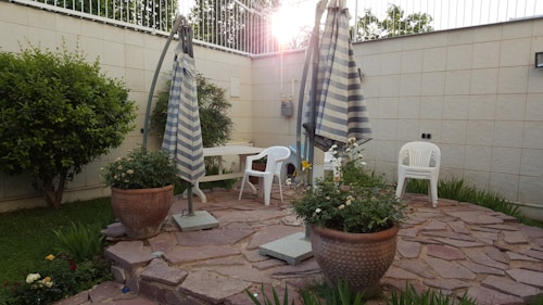 A small patio area features two large striped umbrellas, plastic chairs, and a stone surface. Flower pots with blooming plants add greenery, while a large bush and a tree are visible next to the white wall. The late afternoon sun casts a warm glow, creating shadows on the ground.