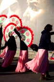 A group of performers dressed in long pink skirts and dark tops holding vibrant fans with red trim. They appear to be dancing or moving in unison. The background features a large mural of a pensive face, adding a dramatic element to the scene.