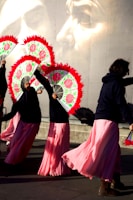 A group of performers dressed in long pink skirts and dark tops holding vibrant fans with red trim. They appear to be dancing or moving in unison. The background features a large mural of a pensive face, adding a dramatic element to the scene.