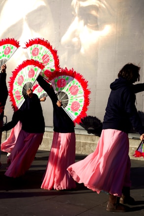 A group of performers dressed in long pink skirts and dark tops holding vibrant fans with red trim. They appear to be dancing or moving in unison. The background features a large mural of a pensive face, adding a dramatic element to the scene.