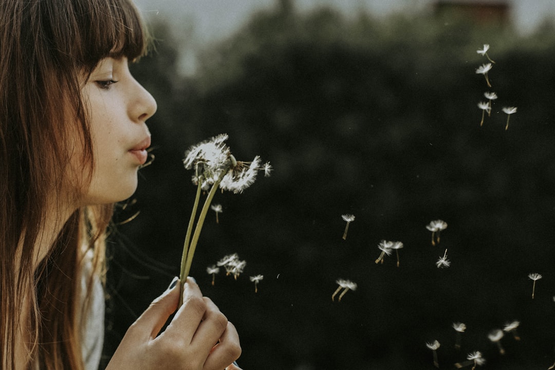 A woman with bangs blows on a dandelion, scattering its seeds
