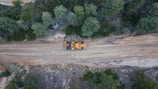 Aerial view of a construction vehicle on a dirt road surrounded by dense green trees. The landscape shows a blend of natural vegetation and a cleared path for machinery, indicating an active construction or forestry area.