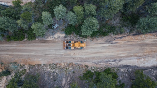 Aerial view of a construction vehicle on a dirt road surrounded by dense green trees. The landscape shows a blend of natural vegetation and a cleared path for machinery, indicating an active construction or forestry area.