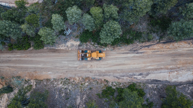 Aerial view of a construction vehicle on a dirt road surrounded by dense green trees. The landscape shows a blend of natural vegetation and a cleared path for machinery, indicating an active construction or forestry area.