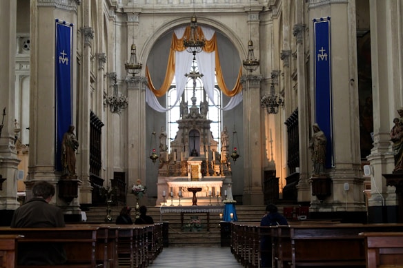 A beautifully decorated church interior with a high ceiling and stone architecture. The center features a detailed altar with religious symbols and candles, framed by tall pillars and arched windows adorned with draped fabric in gold and white. Statues stand along the sides, and chandeliers hang from the ceiling. Several people are seated on wooden pews, facing the altar.