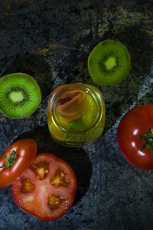 A composition featuring sliced green kiwis and red tomatoes arranged on a textured dark surface. A jar filled with a yellowish liquid and containing a piece of citrus is placed at the center, creating a contrast with the surrounding fruits.