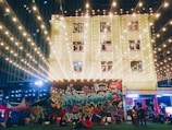 A vibrant scene featuring a building adorned with numerous string lights creating a warm glow. The wall of the building is covered in colorful graffiti art. Several people are gathered in front, some sitting on the grass, which suggests a relaxed, social atmosphere. The background includes additional structures and city lights indicating an urban setting.