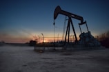 An oil pumpjack is situated in an open area, fenced off and silhouetted against a sunset or sunrise. The sky transitions from deep blue to warm hues near the horizon. The surroundings appear barren and industrial.