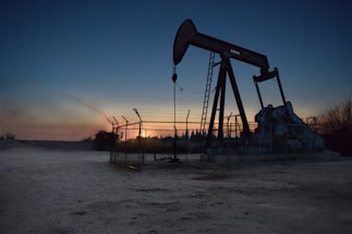 An oil pumpjack is situated in an open area, fenced off and silhouetted against a sunset or sunrise. The sky transitions from deep blue to warm hues near the horizon. The surroundings appear barren and industrial.