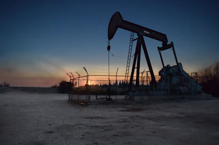 An oil pumpjack is situated in an open area, fenced off and silhouetted against a sunset or sunrise. The sky transitions from deep blue to warm hues near the horizon. The surroundings appear barren and industrial.