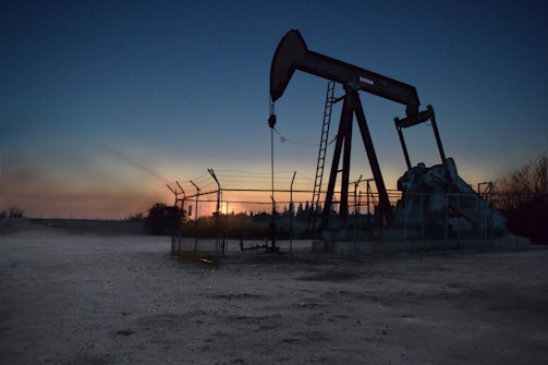 An oil pumpjack is situated in an open area, fenced off and silhouetted against a sunset or sunrise. The sky transitions from deep blue to warm hues near the horizon. The surroundings appear barren and industrial.