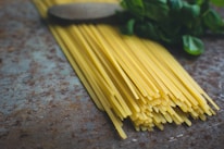 A bundle of uncooked spaghetti pasta lies on a textured surface near some fresh basil leaves. The spaghetti is neatly arranged, and the basil adds a touch of greenery to the composition.