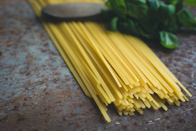 A bundle of uncooked spaghetti pasta lies on a textured surface near some fresh basil leaves. The spaghetti is neatly arranged, and the basil adds a touch of greenery to the composition.