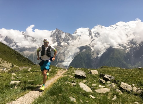 A person is running on a mountain trail surrounded by lush green grass and rugged rocks. In the background, majestic snow-capped mountains are partially covered by white clouds, beneath a clear blue sky. The runner is wearing athletic gear, including a cap and a vest.