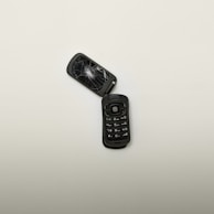 A broken flip phone with a shattered screen lies open on a plain, light-colored surface. The keypad is intact, showing numbers and symbols, while the screen is severely cracked.