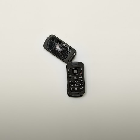 A broken flip phone with a shattered screen lies open on a plain, light-colored surface. The keypad is intact, showing numbers and symbols, while the screen is severely cracked.