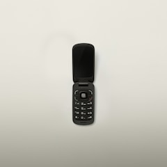 A black flip phone with a compact design is open, displaying its keypad and screen. The phone has a circular navigation button and standard numerical keys. Set against a plain white background.