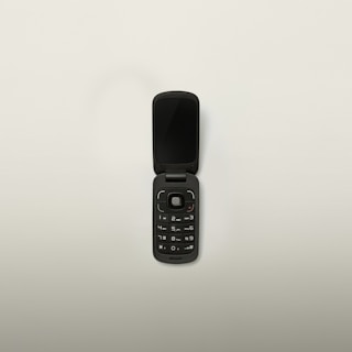 A black flip phone with a compact design is open, displaying its keypad and screen. The phone has a circular navigation button and standard numerical keys. Set against a plain white background.