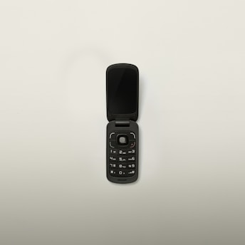 A black flip phone with a compact design is open, displaying its keypad and screen. The phone has a circular navigation button and standard numerical keys. Set against a plain white background.