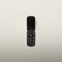 A black flip phone with a compact design is open, displaying its keypad and screen. The phone has a circular navigation button and standard numerical keys. Set against a plain white background.