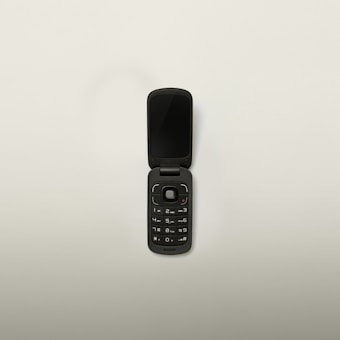 A black flip phone with a compact design is open, displaying its keypad and screen. The phone has a circular navigation button and standard numerical keys. Set against a plain white background.