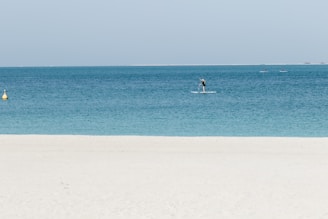 A serene beach scene with clear turquoise water. A person is paddleboarding near the shore, and a few more paddleboarders are visible further out on the water. The sky is clear and light blue, and a yellow buoy can be seen in the water close to the left side.