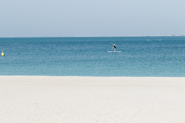 A serene beach scene with clear turquoise water. A person is paddleboarding near the shore, and a few more paddleboarders are visible further out on the water. The sky is clear and light blue, and a yellow buoy can be seen in the water close to the left side.