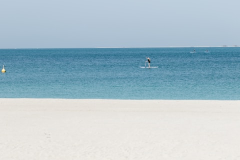 A serene beach scene with clear turquoise water. A person is paddleboarding near the shore, and a few more paddleboarders are visible further out on the water. The sky is clear and light blue, and a yellow buoy can be seen in the water close to the left side.