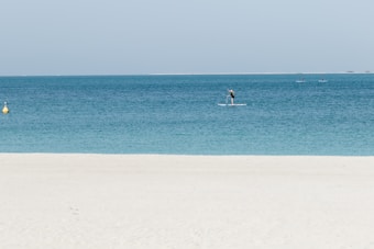 A serene beach scene with clear turquoise water. A person is paddleboarding near the shore, and a few more paddleboarders are visible further out on the water. The sky is clear and light blue, and a yellow buoy can be seen in the water close to the left side.