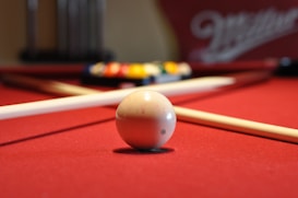 A close-up of a white cue ball on a red pool table with a cue stick positioned nearby. In the background, colorful billiard balls are gathered, slightly out of focus.
