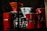 A collection of coffee-making equipment including red and white drippers, a red espresso maker, and glass coffee pots arranged on a shelf. The lighting creates a cozy, intimate atmosphere with shadows and reflections visible on the glass surfaces.