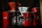 A collection of coffee-making equipment including red and white drippers, a red espresso maker, and glass coffee pots arranged on a shelf. The lighting creates a cozy, intimate atmosphere with shadows and reflections visible on the glass surfaces.