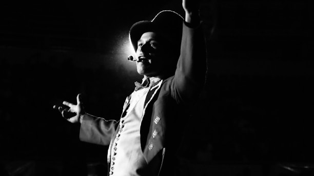 A person in a dramatic pose is highlighted against a dark background, wearing a top hat and holding a microphone close to their mouth. The lighting casts a spotlight effect, creating strong contrasts and shadows.
