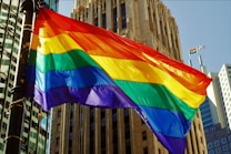 A vibrant rainbow flag waves prominently against a backdrop of tall city buildings, reflecting sunlight and casting dynamic shadows. The architecture features modern and classic elements, adding depth to the urban scene.