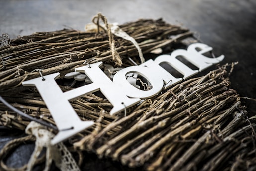 A decorative piece made of intertwined twigs and branches with the word 'Home' prominently displayed in white letters on top. The arrangement conveys a rustic and natural aesthetic, accented by textures of wood and subtle shadows.